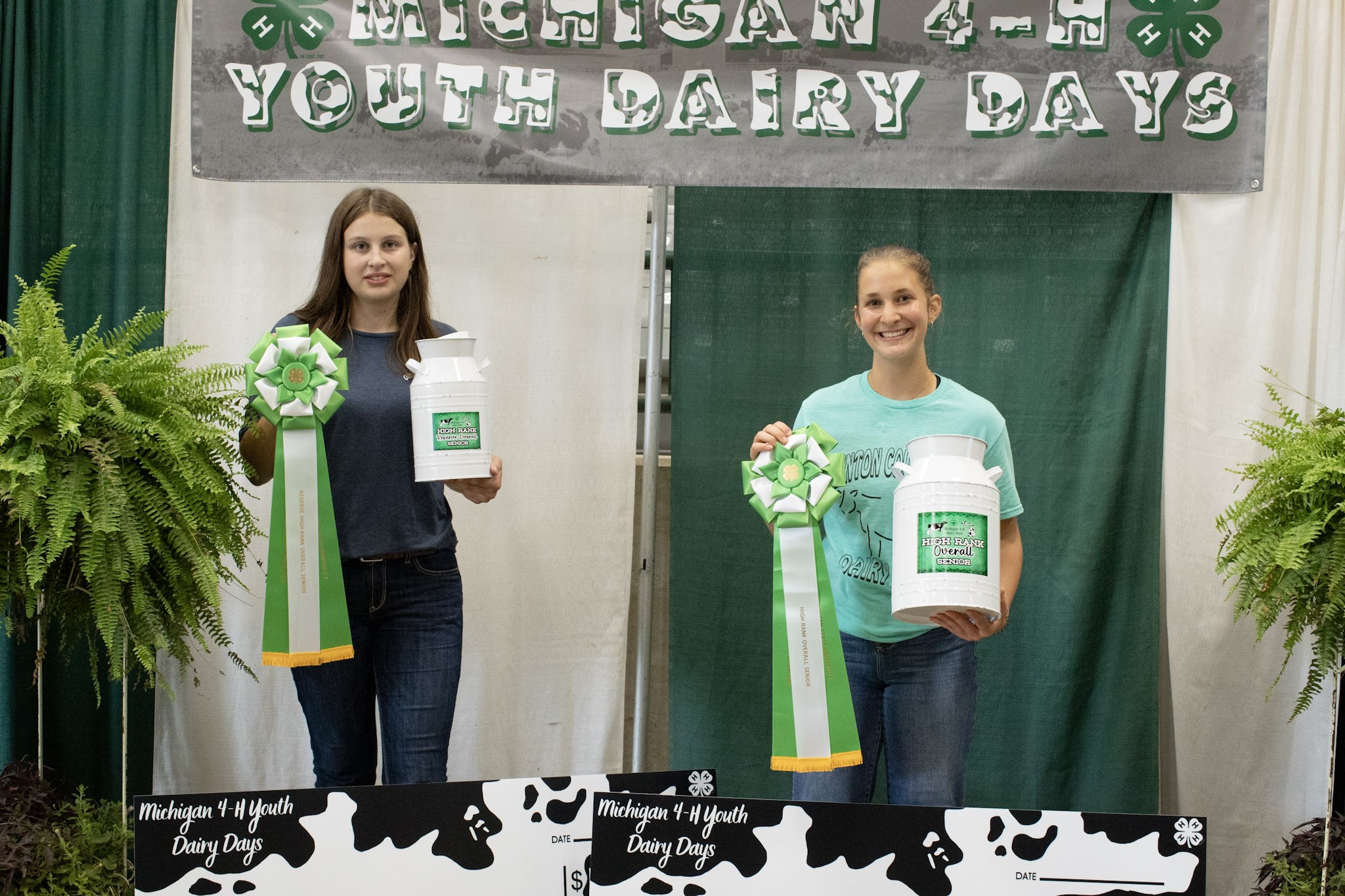 Two young ladies holding up prizes and ribbons in front of a Michigan 4-H Youth Dairy Days banner.